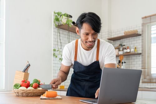 Man in kitchen looking at recipes on laptop while cooking